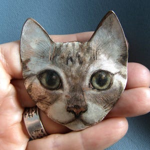 May include: A metal brooch shaped like a cat head with etched fur details in shades of silver, brown, and white. The cat features large, dark, expressive eyes and pointed ears, with a metallic finish that highlights the textured engraved lines.