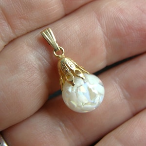 May include: A gold-colored pendant with a pear-shaped, iridescent white stone. The pendant has a decorative filigree cap and a small loop for a chain. The stone appears to be made of multiple pieces.
