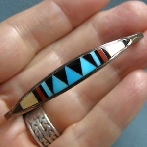 May include: A slender, elongated silver bar brooch featuring a geometric inlay pattern. The design consists of alternating triangular and rectangular segments in shades of turquoise, black, white, and red, set within a polished silver frame.