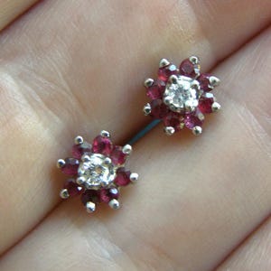 May include: A silver barbell-style body piercing jewelry piece. Each end features a flower design with a central clear stone surrounded by smaller, deep red stones. The jewelry is held in a hand.