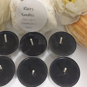 May include: Six black tea light candles with white wicks. The image includes a seashell, white flowers, and a label that reads "Klairs Kandles Tea Lights Handmade in Wales". Candles are on a white surface.