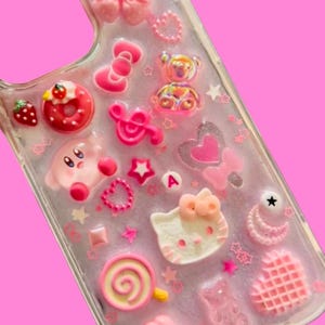 May include: A clear phone case adorned with pink and white charms. The charms include a bow, a cross, a Hello Kitty face, a Kirby character, a donut, a strawberry, stars, hearts, and a lollipop. The case is set against a pink backdrop.