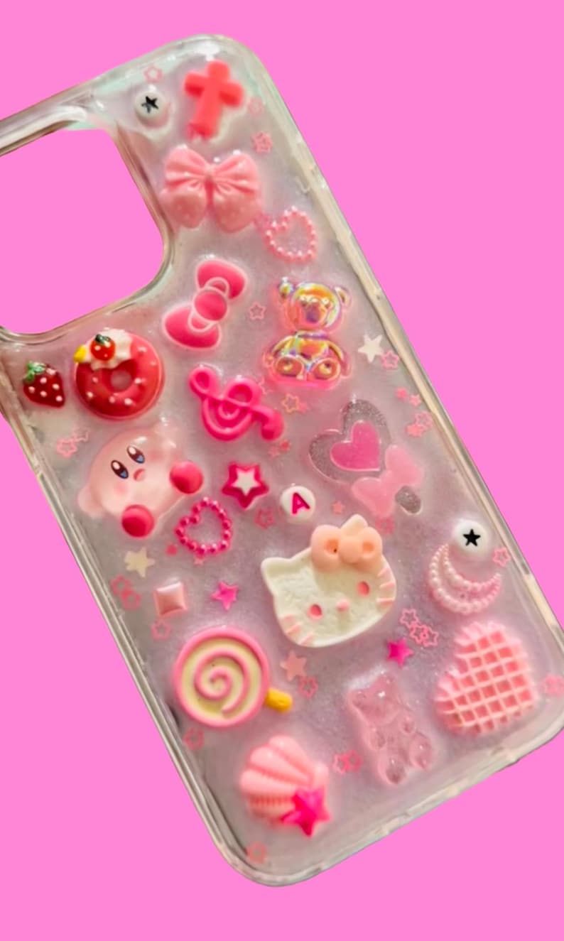 May include: A clear phone case adorned with pink and white charms. The charms include a bow, a cross, a Hello Kitty face, a Kirby character, a donut, a strawberry, stars, hearts, and a lollipop. The case is set against a pink backdrop.