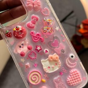 May include: Clear phone case decorated with pink charms, including a cross, bows, a Hello Kitty face, a Kirby character, a donut, and a lollipop. The case is adorned with stars, hearts, and other decorative elements.