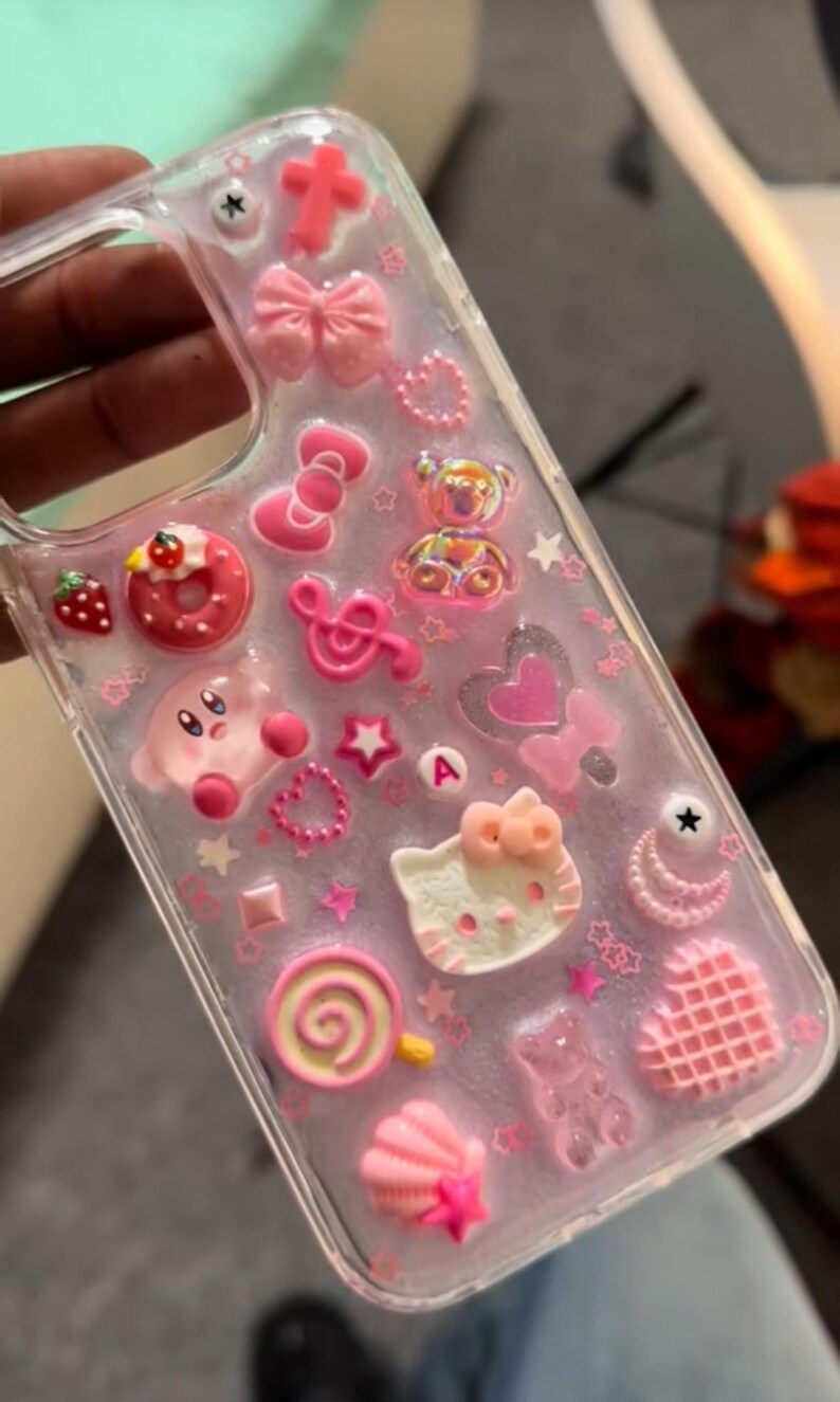 May include: Clear phone case decorated with pink charms, including a cross, bows, a Hello Kitty face, a Kirby character, a donut, and a lollipop. The case is adorned with stars, hearts, and other decorative elements.