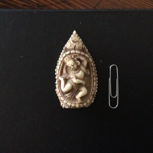 May include: A carved, off-white figurine of a dancing figure within an oval frame. The figurine is placed on a black surface next to a silver paperclip. The carving has a detailed, textured appearance.
