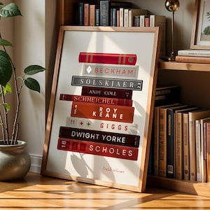 May include: A framed print featuring a stack of books with names like Beckham, Solskjær, and Schmeichel. The print is displayed on a wooden bookshelf, alongside other books and a framed botanical print. A potted plant sits nearby.