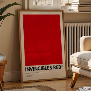 May include: A rectangular framed wall art print featuring a solid bright red background with the text INVINCIBLES RED, Arsenal Ar-1886, and #db0007 printed in black at the bottom. The print is displayed in a light wood frame resting on the floor.