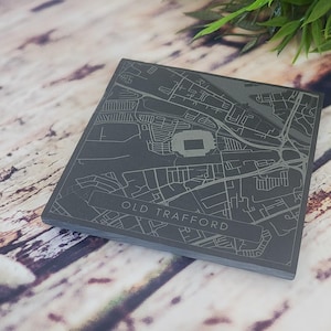 May include: A dark grey square coaster featuring a detailed map of the Old Trafford area, including the stadium. The map is etched into the coaster's surface. The text "OLD TRAFFORD" is visible.