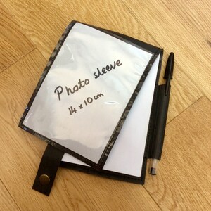 May include: A black photo sleeve with a clear plastic front, measuring 14 x 10 cm. The text "Photo sleeve" is handwritten on the front. A black pen is in a matching pen holder attached to the sleeve.