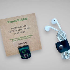 May include: A black leather cable tidy with a silver button, handmade from 100% recycled rubber inner tubes. The tidy is holding a pair of white earbuds. The packaging includes the text "Planet Rubber", "Handmade from 100% recycled rubber inner tubes", "www.etsy.com/shop/planetrubber", "planetrubber@outlook.com", and "Cable tidy".