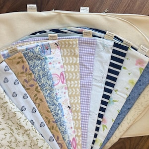 May include: A selection of patterned fabrics, including floral, striped, and gingham designs, arranged in a fan shape. The fabrics are displayed against a cream-coloured oval background. Each fabric piece has a small tag attached.
