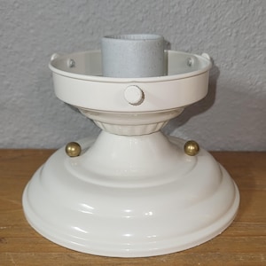 Può includere: Un lampadario bianco con un portalampada centrale e una base decorativa. L'apparecchio ha una finitura lucida, dettagli color ottone e un design classico. Il lampadario è progettato per essere montato a soffitto.