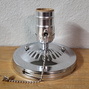 Puede incluir: Una lámpara de techo cromada con una cadena de tracción. La lámpara tiene una base redonda con un patrón decorativo y tres pequeños acentos redondos. El portalámparas es visible en la parte superior.