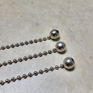 May include: Close-up of three silver ball chains. Each chain has small, evenly spaced ball links, with a larger ball at the end. The chains are on a light-colored surface.