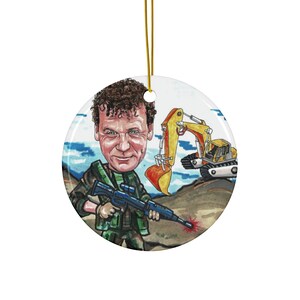 May include: Round ceramic ornament featuring a cartoon illustration of a man holding a rifle, with an excavator in the background. The man has curly brown hair and is wearing a green military-style outfit. The ornament has a gold string for hanging.