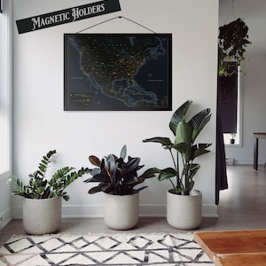 May include: A black framed map of the United States with a dark blue background and yellow and green dots. The map is hanging on a white wall with a sign that reads "Magnetic Holders". There are three potted plants in the room, one with dark green leaves, one with light green leaves, and one with large, dark green leaves. The plants are in grey pots. There is a brown wooden table in the foreground.