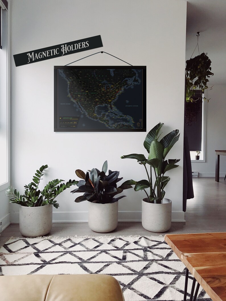 May include: A black framed map of the United States with a dark blue background and yellow and green dots. The map is hanging on a white wall with a sign that reads "Magnetic Holders". There are three potted plants in the room, one with dark green leaves, one with light green leaves, and one with large, dark green leaves. The plants are in grey pots. There is a brown wooden table in the foreground.