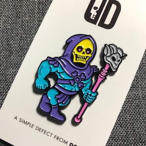 May include: A purple and blue enamel pin of Skeletor from Masters of the Universe. Skeletor is holding a pink staff with a skull on top. The pin is on a white card with the text "A simple defect from DCAY" and a logo of a skull with the letters "DCAY".