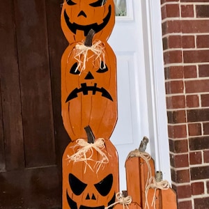 May include: A vertical stack of three orange wooden pumpkins with black painted jack-o'-lantern faces and raffia bows, accompanied by two smaller, plain orange wooden pumpkins with twine bows, all featuring dark brown painted stems.