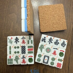 May include: Four square coasters crafted from white Mahjong tiles with blue sides and cork backings. Two coasters are face-up, displaying various Mahjong characters, symbols, and Joker tiles in red, green, and black ink. One coaster is flipped to show the cork base.