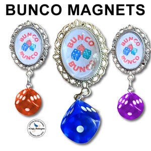 May include: Three oval magnets with silver filigree frames featuring the text BUNCO and illustrations of dice. Each magnet has a dangling translucent die in orange, blue, or purple. The text BUNCO MAGNETS appears at the top of the image.