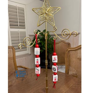 Pode incluir: Um suporte de exibi&ccedil;&atilde;o de metal dourado com um topo em forma de estrela, segurando duas fileiras de pe&ccedil;as decorativas de Mahjong. As pe&ccedil;as s&atilde;o brancas com marca&ccedil;&otilde;es vermelhas e pretas, e contas e borlas vermelhas.