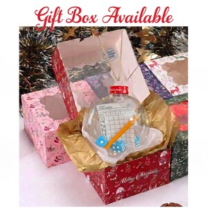 May include: A clear, spherical Christmas ornament containing a Bunco score sheet, a yellow pencil, and two blue dice. The ornament has a red cap and is nestled in gold tissue paper inside a red box with white holiday patterns and text.