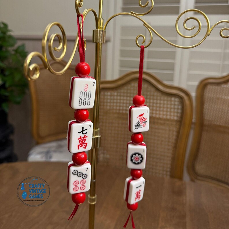 Pode incluir: Dois ornamentos decorativos de pe&ccedil;as de Mahjong. Cada ornamento apresenta pe&ccedil;as brancas com detalhes em vermelho, amarradas com contas e fita vermelhas. Um ornamento exibe o texto "七萬" e o outro uma ilustra&ccedil;&atilde;o de um p&aacute;ssaro.
