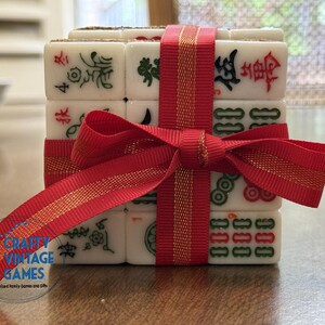 May include: A stack of white rectangular mahjong tiles featuring engraved characters and symbols in red, green, and black ink. The tiles are bound together by a bright red ribbon tied in a bow. A circular logo with the text Crafty Vintage Games is visible.