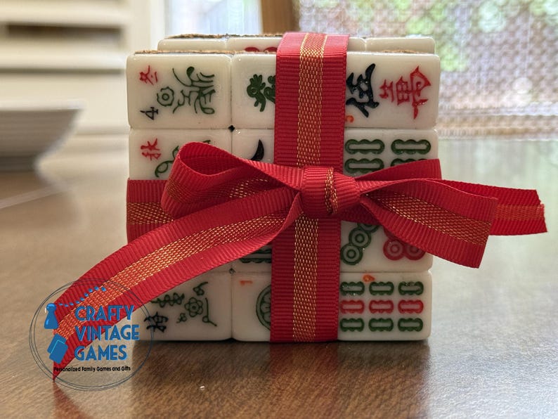 May include: A stack of white rectangular mahjong tiles featuring engraved characters and symbols in red, green, and black ink. The tiles are bound together by a bright red ribbon tied in a bow. A circular logo with the text Crafty Vintage Games is visible.