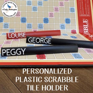 May include: Three personalised plastic Scrabble tile holders in red and black. The red holder features the name LOUISE in white text, while the two black holders display the names GEORGE and PEGGY in white text, positioned on a game board.