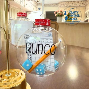 May include: A clear, spherical Christmas ornament containing a Bunco score sheet, a yellow pencil, and two light blue dice. The ornament features a red cap with the word Christmas printed in white. The word BUNCO is printed in black on the front.