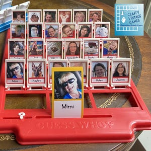 Może przedstawiać: Czerwono-biała plansza do gry Guess Who? z r&oacute;żnymi zdjęciami os&oacute;b i psa. Plansza do gry ma mechanizm przesuwny do ujawniania zdjęć. Plansza do gry znajduje się na drewnianym stole.