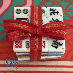 May include: A stack of square white Mahjong tiles featuring various black, red, and green symbols and characters, including a red Joker tile. The tiles are bound together with a red ribbon tied in a bow, featuring a thin gold stripe down the center.