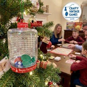 May include: A clear plastic Christmas ornament shaped like a jar, featuring a printed Sequence game board on the exterior, a red lid with Merry Christmas text, and green and red game chips inside, hanging from a green string on a pine tree.