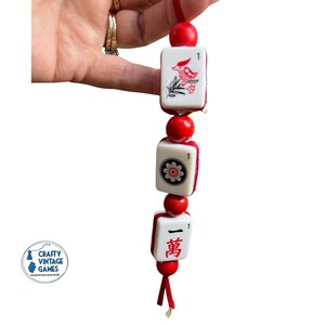 Pode incluir: Uma sequ&ecirc;ncia de tr&ecirc;s pe&ccedil;as de Mahjong vintage com detalhes e contas vermelhas. As pe&ccedil;as apresentam s&iacute;mbolos e ilustra&ccedil;&otilde;es vermelhas e pretas, incluindo um p&aacute;ssaro e uma flor. As pe&ccedil;as s&atilde;o amarradas em um cord&atilde;o vermelho.