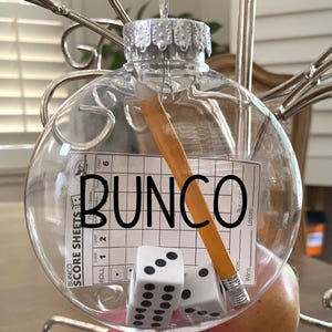 May include: A clear, spherical glass ornament containing a miniature yellow pencil, two white six-sided dice with black pips, and a small Bunco score sheet. The word BUNCO is printed in large black capital letters across the front of the ornament.
