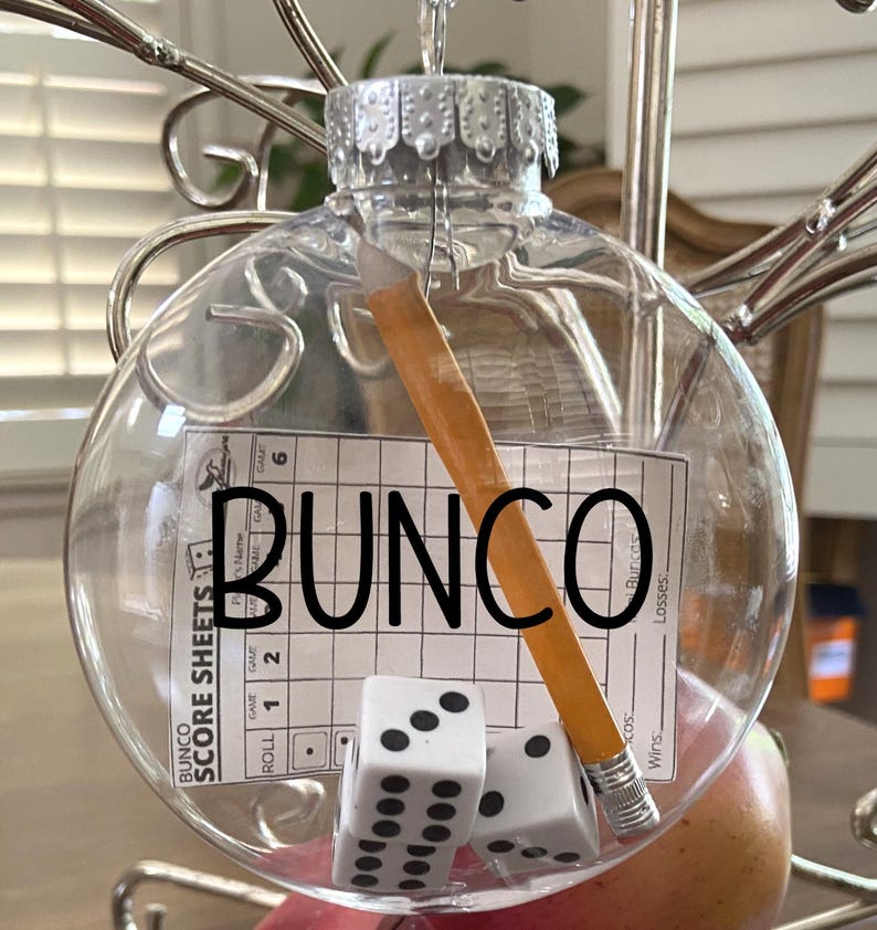 May include: A clear, spherical glass ornament containing a miniature yellow pencil, two white six-sided dice with black pips, and a small Bunco score sheet. The word BUNCO is printed in large black capital letters across the front of the ornament.