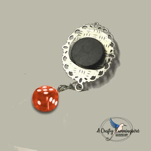 May include: A decorative magnet featuring a silver-toned oval frame with an intricate filigree border and a black circular magnetic backing. A small, translucent red six-sided die with white pips hangs from the frame by a silver metal jump ring.