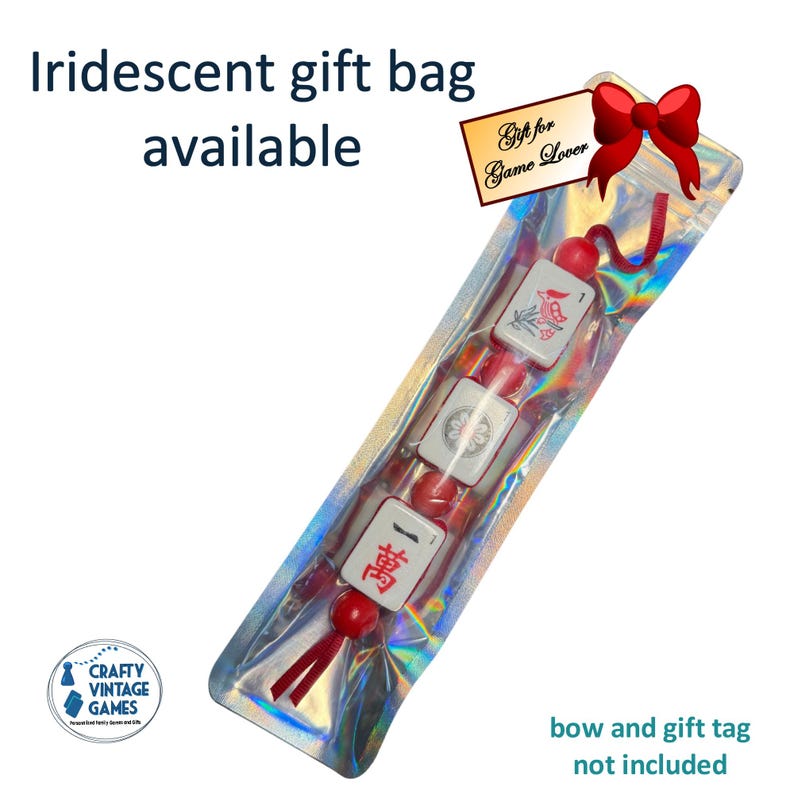 Pode incluir: Uma sacola de presente iridescente transparente contendo uma sequ&ecirc;ncia de pe&ccedil;as de Mahjong brancas e vermelhas. As pe&ccedil;as t&ecirc;m caracteres e s&iacute;mbolos vermelhos. A sacola tem uma etiqueta que diz "Gift for Game Lover". O texto "Iridescent gift bag available" tamb&eacute;m &eacute; vis&iacute;vel.