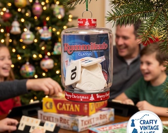 May include: A clear plastic ornament filled with white Rummikub game tiles. The container features a red ribbon printed with the text Merry Christmas and a small tree icon. The ornament is suspended by a green string from a pine branch.