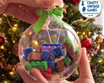 May include: A clear, spherical plastic Christmas ornament filled with various colorful plastic building blocks. The ornament is topped with a green ribbon tied in a bow, featuring white text that reads Merry Christmas, and has a silver metal hanger.