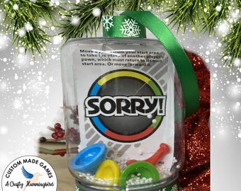 May include: A clear plastic ornament filled with small white beads, containing Sorry! game pieces in blue, yellow, red, and green. The ornament features a green ribbon with white snowflake patterns and a card with game instructions.