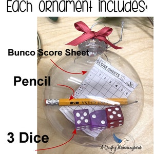 May include: A clear, spherical glass ornament topped with a red ribbon bow. Inside the ornament are a Bunco score sheet, a yellow pencil with a metal ferrule and eraser, and three six-sided dice in purple, light purple, and red colors.