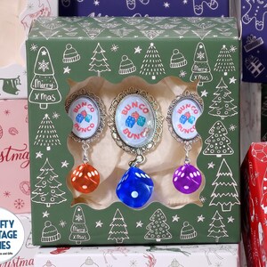 May include: A green cardboard box with a window displaying three Bunco game markers. Each marker features an oval metal frame with the text Bunco and a graphic of three dice, with a dangling translucent orange, blue, or purple die attached.