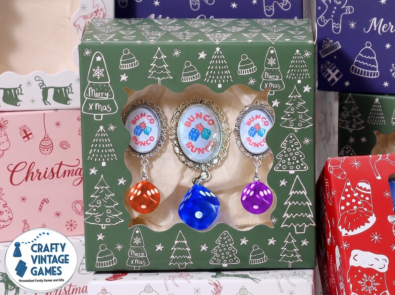 May include: A green cardboard box with a window displaying three Bunco game markers. Each marker features an oval metal frame with the text Bunco and a graphic of three dice, with a dangling translucent orange, blue, or purple die attached.