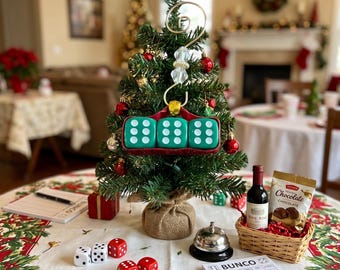 May include: A small artificial Christmas tree decorated with red ornaments and a hanging ornament featuring three green dice. The table includes a small wicker basket with a miniature wine bottle and chocolate, a silver bell, dice, and Bunco score pads.