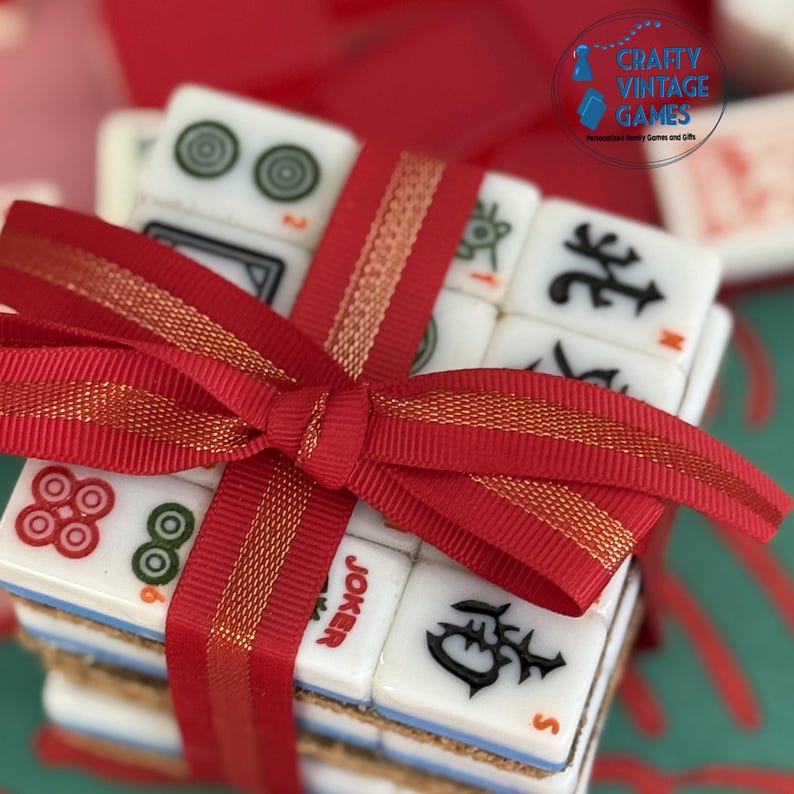 May include: A stack of white Mahjong tiles with black, red, and green symbols, including a Joker tile, tied together with a red ribbon featuring a gold metallic edge. The tiles have a light blue base and a cork backing, arranged in a square formation.