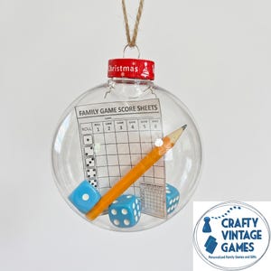 May include: A clear spherical Christmas ornament containing a printed family game score sheet, a yellow wooden pencil, and three light blue dice with white pips. The ornament has a red cap with the word Christmas and a small tree icon printed on it.